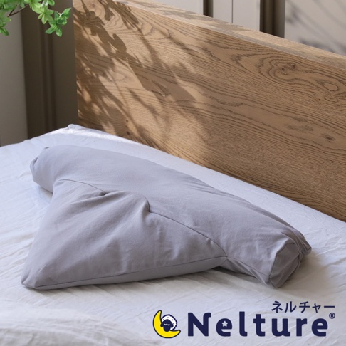 一個淺灰色的長條形枕頭，表面光滑，一端呈現三角形，另一端較為圓潤，枕頭置於白色床單上，背景為木質床頭板。