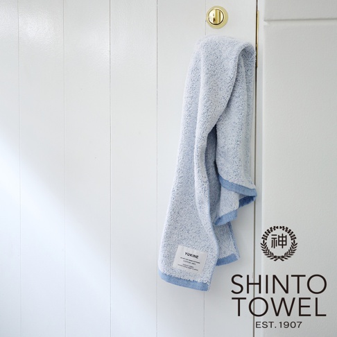 YUKINE 雪中漫步迷你浴巾，淺藍色，蓬厚柔軟的有機棉材質，帶有藍色滾邊，懸掛於白色木紋牆面。