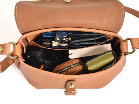 棕色皮革馬鞍包，內部裝有藍色錢包、鑰匙包、黑色相機、書本和綠色小皮夾。包款配有黃銅色金屬扣環和可調節肩帶。