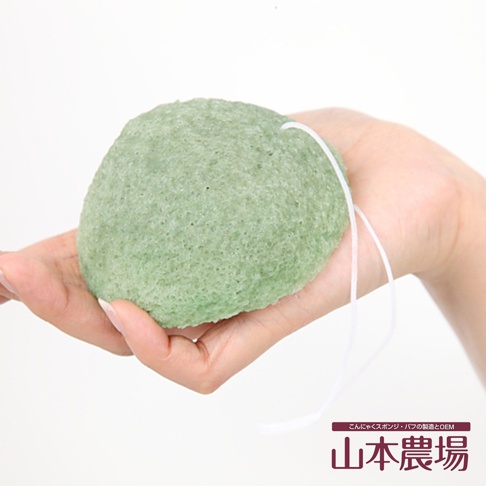 手持一顆圓形、淺綠色的蒟蒻海綿，表面有細密的孔洞，一條白色細繩穿過海綿。