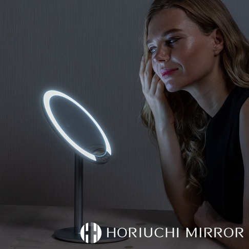 桌上型LED化妝鏡，圓形鏡面，邊緣環繞著明亮的LED燈光，鏡架與底座為銀灰色金屬材質，一位女士正在鏡前化妝。