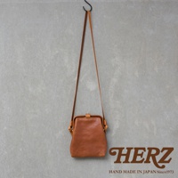 職人手作皮革Organ迷你杜勒斯側背包，棕色皮革，搭扣設計，附有細長肩帶，掛在灰色牆壁上。