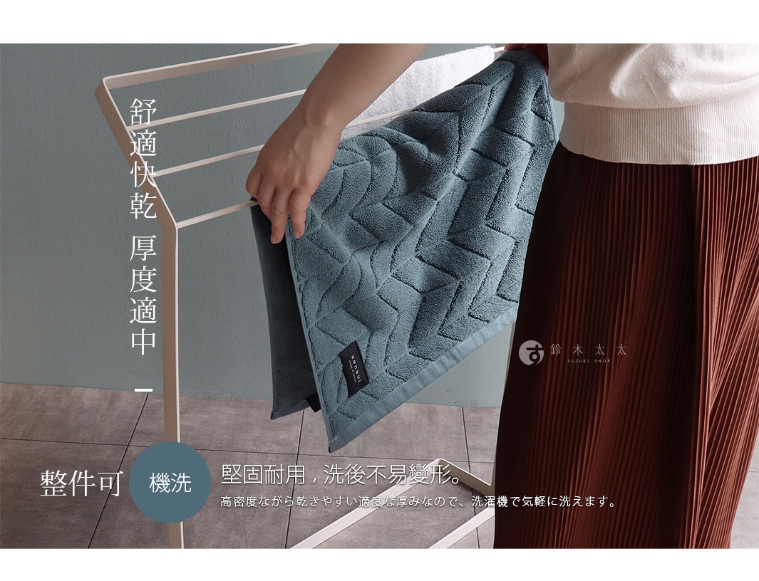 一件藍綠色的毛巾被一隻手拿著，毛巾上有菱形圖案，背景是簡約的白色掛架，下方有文字說明其材質和耐用性。