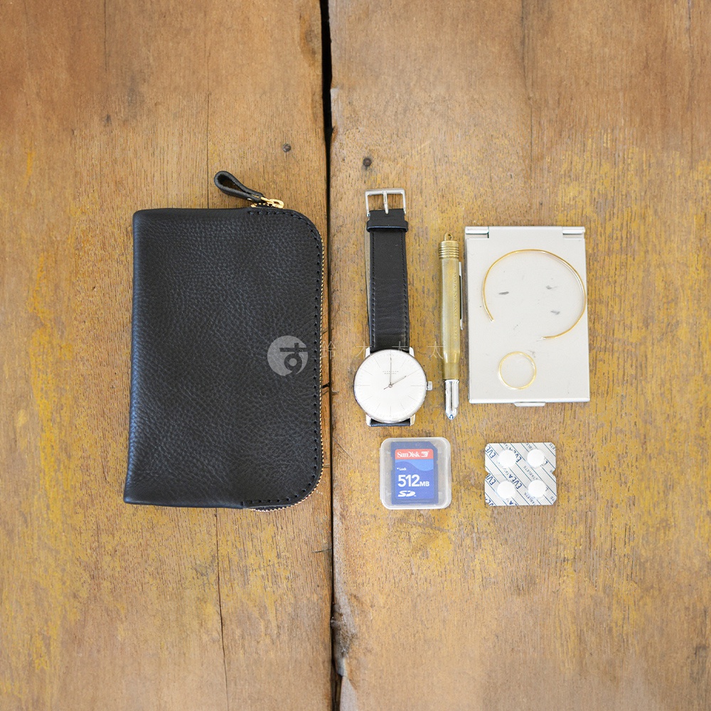 黑色皮革雙拉鍊收納袋，搭配金色拉鍊頭與品牌標誌。旁邊擺放一只簡約白色錶盤的黑色錶帶手錶、一支金色鋼筆、一個銀色方形小鏡盒，內有金屬細圈飾品，下方還有一個 SanDisk 512MB 記憶卡和兩片白色藥片包裝。