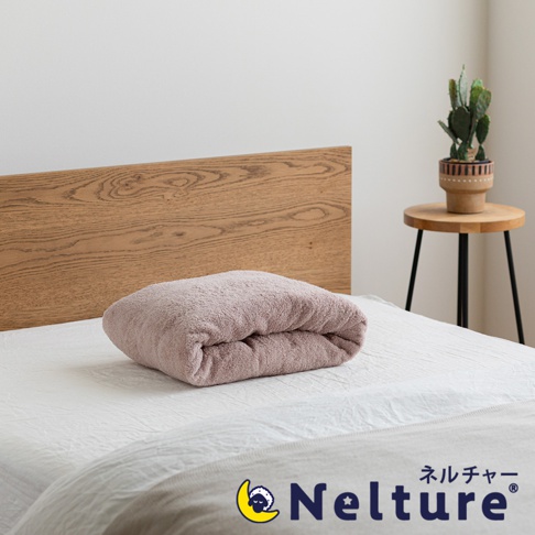 一條粉色毛巾狀的枕頭，以甜甜圈形狀對折，放在白色床單上，背景是木紋床頭板和裝有仙人掌的圓形邊桌。