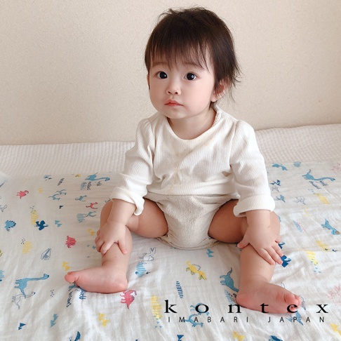 一名嬰兒身穿白色長袖上衣與米色尿布，坐在印有恐龍、兔子等童趣塗鴉圖案的二層紗嬰兒毯上。