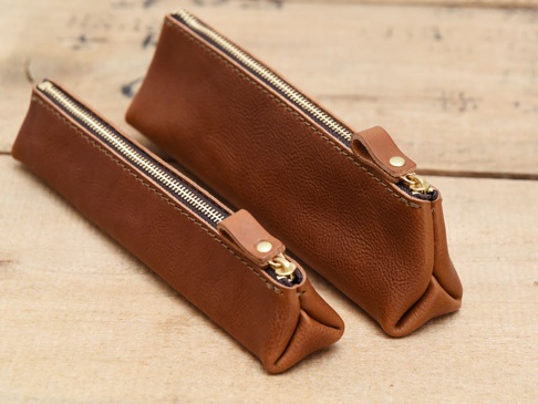 兩款棕色皮革製成的三角收納筆袋，帶有黃銅色拉鍊和皮革拉環，置於淺色木質背景上。