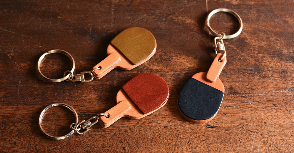 職人手作皮革桌球拍造型鑰匙圈，包含棕色、紅色和黑色等款式，皆以皮革材質製作，搭配金屬鑰匙圈。
