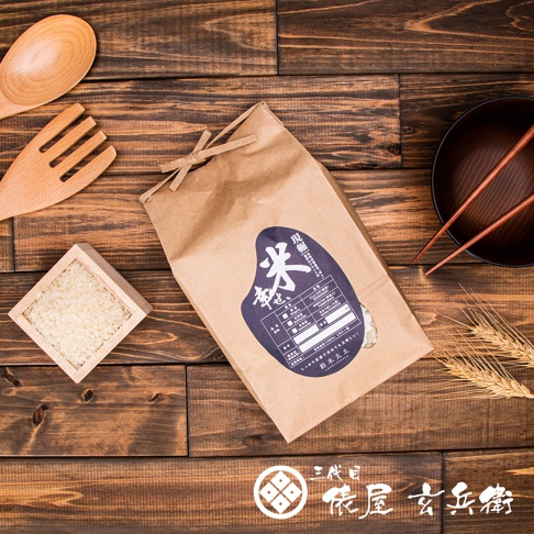 淺棕色紙袋裝著北海道夢美人米，上面貼有深藍色標籤，標籤上印有日文文字及產品資訊。一旁有木質小方盒裝著少量米粒，以及木製湯勺和叉子。背景為木紋桌面，右側有深色碗和兩根紅色筷子，以及兩支麥穗。
