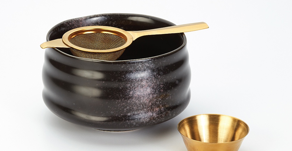 一個帶有金色網狀濾網的黑色陶瓷茶碗，茶碗表面有細小的斑點紋理，旁邊有一個小巧的金色金屬杯。