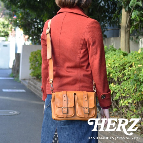 一位女性背著日本HERZ品牌職人手作的淺棕色皮革斜背包，包款設計有兩個前口袋與皮帶扣環，呈現復古質感，背景為戶外街道與綠色植栽。