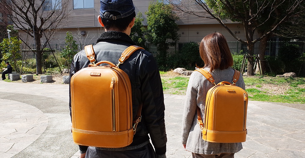 兩名人士背著職人手作皮革立體商務後背包，後背包為淺棕色皮革材質，設計簡潔，帶有拉鍊和提把。