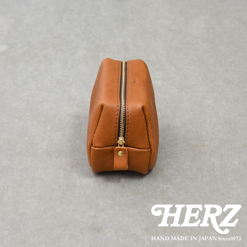 側面視角的日本HERZ職人手作皮革收納包，呈現經典暖棕色質感與細緻的縫線工藝，配有黃銅色金屬拉鍊與皮革拉環，下方印有白色HERZ與手工製作說明字樣，整體風格簡約且具有溫潤的手作感。