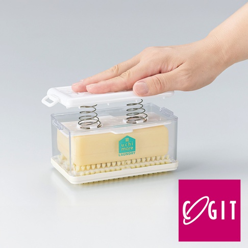 透明塑膠盒內裝有黃色洗衣皂，下方為白色底座與黃色刷毛。盒蓋為白色，內有兩根彈簧，一隻手正將盒蓋壓下。