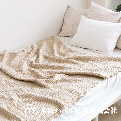 米色三層紗波紋毯鋪展在床上，質地輕透，表面有細緻的波浪紋理。毯子搭在白色床單上，旁邊擺放著淺色和白色的抱枕。