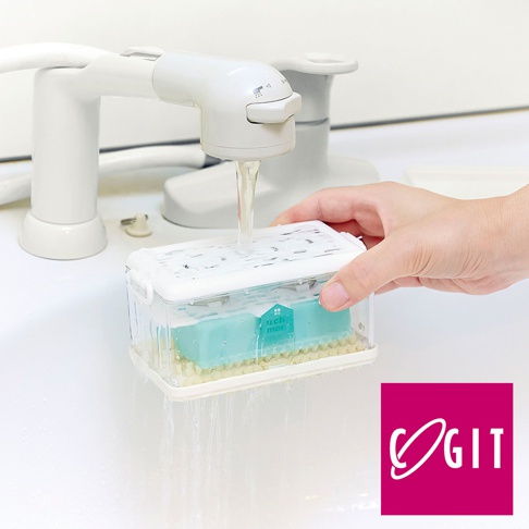 透明塑膠盒內裝有一塊淺藍色洗衣皂，下方有黃色刷毛。水龍頭正將水注入盒中，水流從盒底和側邊的縫隙流出。一隻手正拿著盒子。