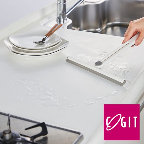 白色的刮水器，帶有銀色旋轉接頭，正在擦拭有水漬的白色檯面。旁邊疊著幾個白色盤子，上面放著帶木質手柄的叉子和勺子。