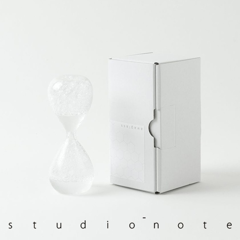 awaglass 泡時計，由透明玻璃製成，中間收窄，上下兩端為球狀。頂部球體內充滿白色泡沫，底部球體內也聚集了白色泡沫。產品旁有一個白色的紙盒，盒身上印有「awaglass」字樣和蜂巢圖案。