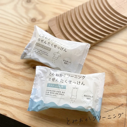 兩塊白色包裝的洗衣皂，上面印有日文說明和衣物圖示，置於淺色木紋背景上，背景後方可見一個木製的洗滌板。