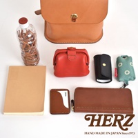 職人手作皮革翻蓋迷你後背包，搭配紅色零錢包、黑色鑰匙包、綠色環保袋、棕色卡夾、長夾及一本筆記本，與一瓶裝滿皮革製品的透明瓶子。