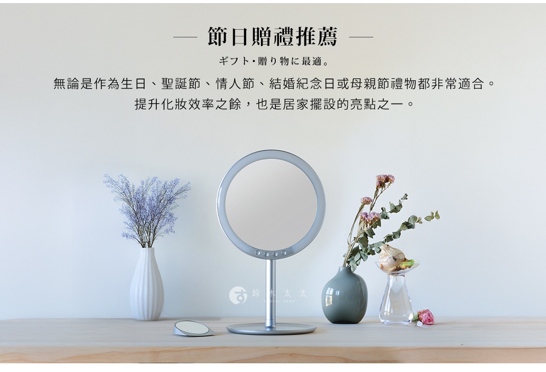 銀色金屬材質的桌上型化妝鏡，圓形鏡面外圍有LED燈帶，鏡座為圓形底座。鏡子前方有三個圓形按鈕。右側有一束乾燥花，左側有一束紫色小碎花，兩束花分別置於白色和灰綠色花瓶中。