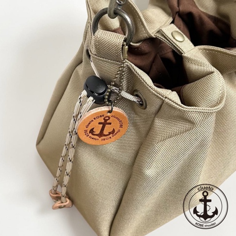 神戶clueto品牌的BALON mini防潑水手提包局部特寫，米色帆布材質包身搭配棕色內襯，包口處設有抽繩與黑色調節扣，並懸掛一個印有錨圖案與clueto KOBE字樣的圓形皮革吊飾，整體呈現簡約休閒風格。