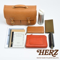 一個棕色皮革的兩用單肩包，配有兩個插扣和提把。周圍散落著一本筆記本、一個長夾、一個橘紅色皮夾、一個綠色長夾、一個黑色筆袋和一個銀色保溫瓶。