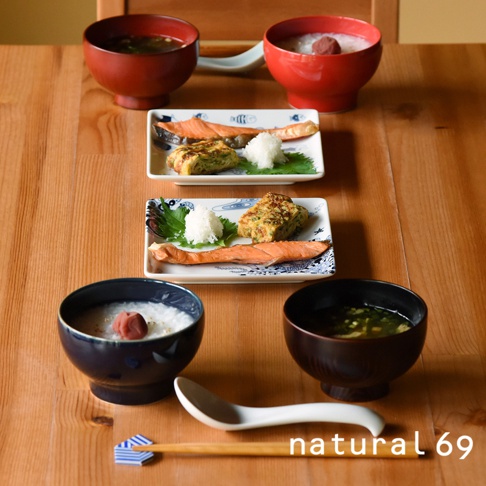 兩個白色方形餐盤，各盛有烤鮭魚、日式煎蛋捲、蘿蔔泥及紫蘇葉，擺放在木製桌面上，上方有兩個紅色湯碗。