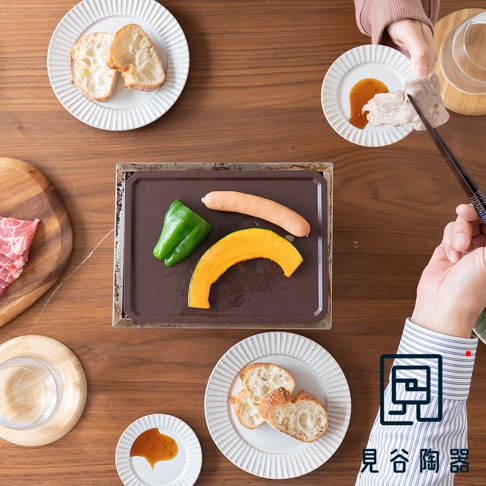 長方形的HAI減煙火山灰烤盤，盤中放有綠色的甜椒、橘黃色的南瓜塊和一根香腸，烤盤旁有切片麵包、肉塊和醬料小碟。