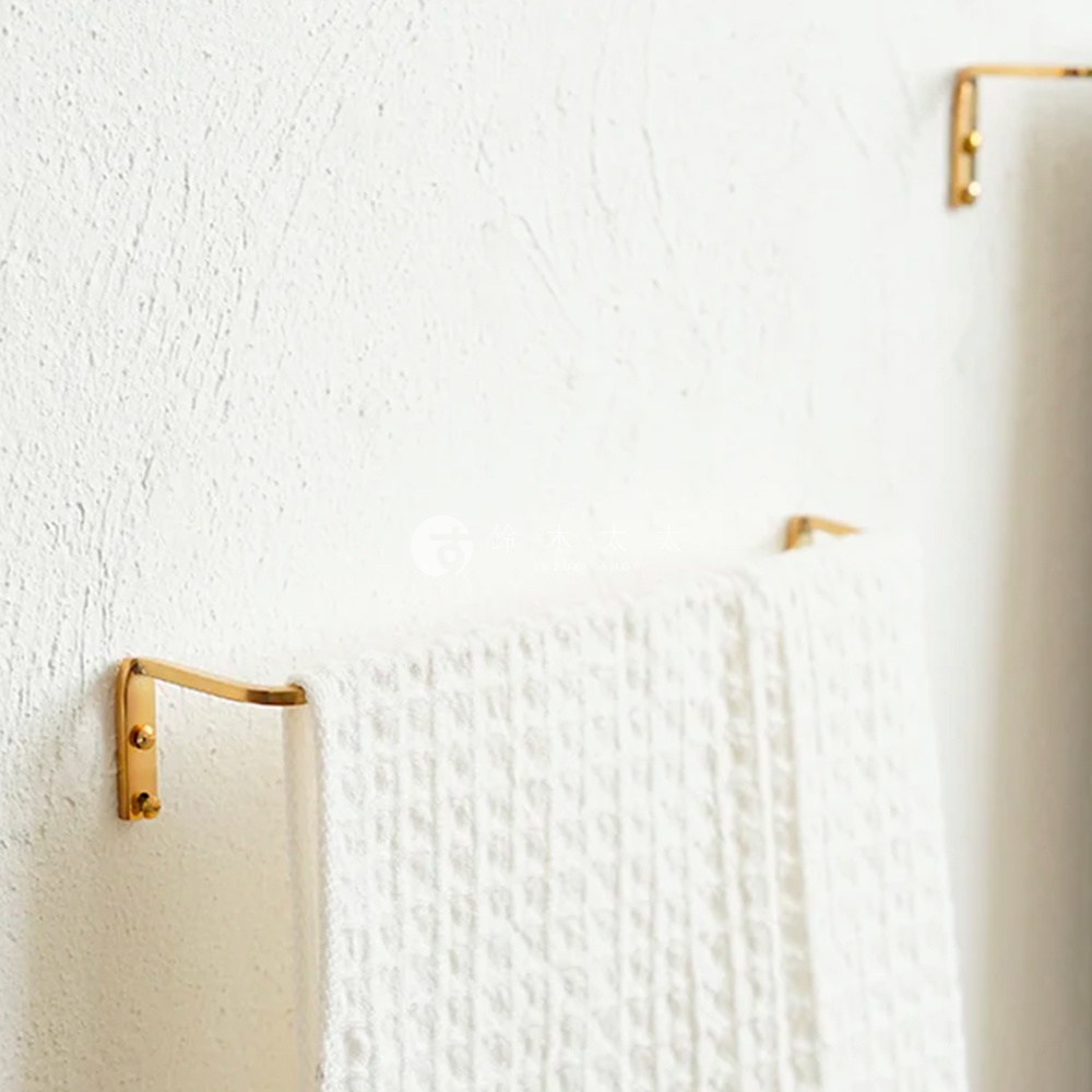 黃銅單桿毛巾架，固定於白色紋理牆壁上，毛巾架呈金色，一條白色紋理毛巾懸掛於其上。