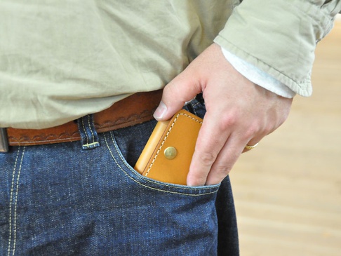 職人手作皮革箱型收納零錢包 (KK-23)，採用天然皮革製作，呈現溫潤的焦糖色調，表面有精緻縫線。零錢包以箱型設計，正面設有金屬按扣，由手部放入牛仔褲口袋中。