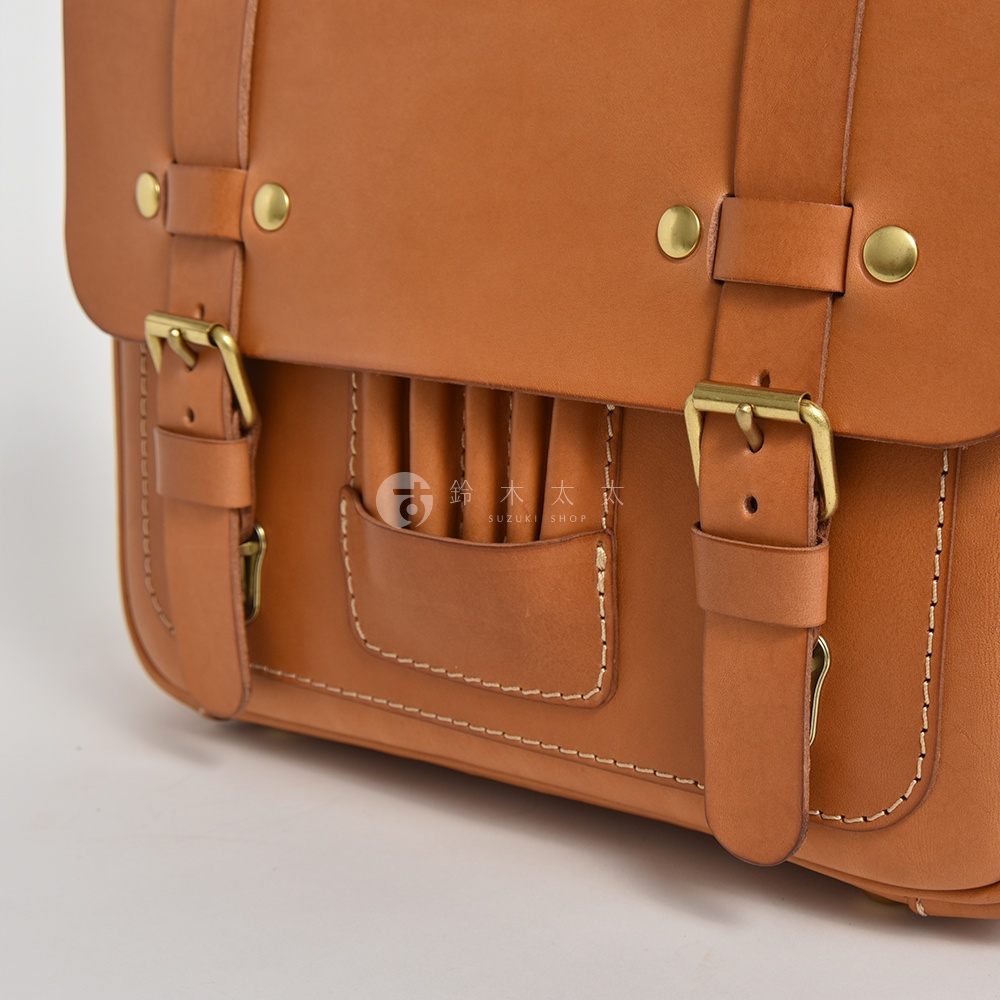 職人手作皮革歐風三用學生包，採用淺棕色皮革，配有黃銅色金屬扣環與鉚釘，包身縫線明顯，前袋設計有褶皺和插袋。
