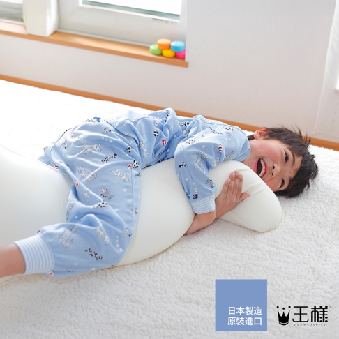 一個男孩穿著印有可愛動物圖案的藍色睡衣，抱著一個白色、柔軟的長形抱枕，側躺在鋪著白色絨毛毯的地板上。