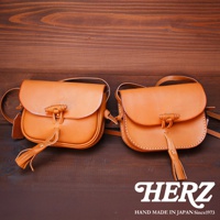 兩款職人手作皮革流蘇側背包，以天然皮革製作，呈現溫潤的焦糖色澤。包包設計簡約，翻蓋處以皮革繩結固定，下方裝飾著皮革流蘇，展現細膩的工藝。