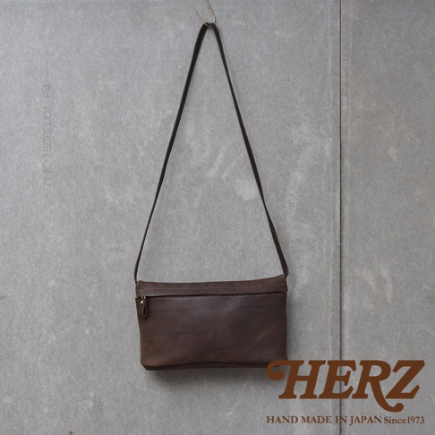 深棕色皮革製成的長方形斜背包，懸掛在灰色水泥牆面上，包包上有拉鍊開口，右下角有HERZ品牌標誌。