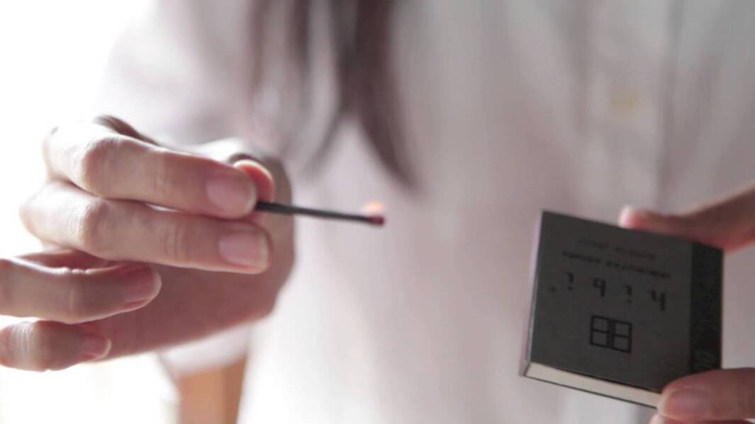 一雙手持著一根點燃的深色火柴，火柴頭有小火焰，旁邊另一隻手拿著一個深灰色小紙盒，紙盒上印有白色文字及圖案。