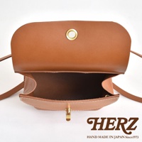 職人手作皮革翻蓋迷你後背包，採用棕褐色真皮製作，包身結構紮實，翻蓋處有金色圓形金屬扣眼，包口處為金色鎖扣，細緻的白色縫線展現手工質感。