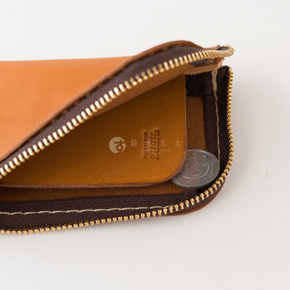 這是一款日本HERZ職人手作的棕色皮革迷你零錢包，呈現開口拉鍊細節，包內印有HERZ品牌標誌與Made in Japan字樣，角落處放置著一枚硬幣作為比例對照，展現細緻的車縫工藝與皮革質感。