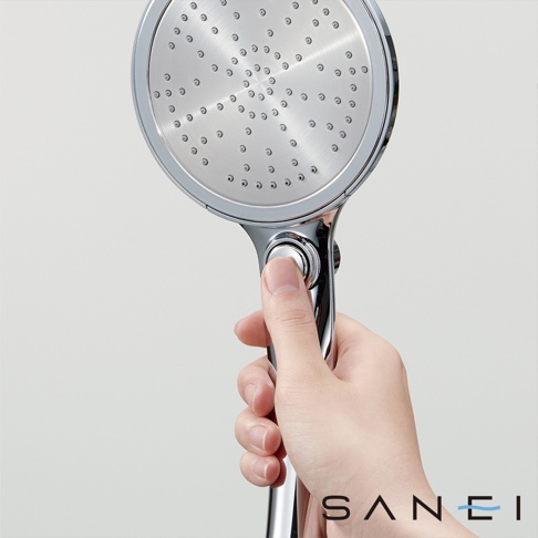 這是一張淺灰色背景的照片，一隻手握著一支三榮品牌的鍍鉻金屬色淋浴蓮蓬頭。蓮蓬頭呈現圓盤狀，面板上有許多細密的出水孔設計，手把處設有一個圓形的開關按鈕，整體外觀簡約且具現代感。