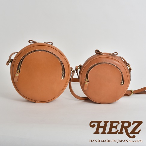 兩款職人手作皮革小圓包，皆為圓形設計，淺棕色皮革材質，配有拉鍊開口及可調節背帶。
