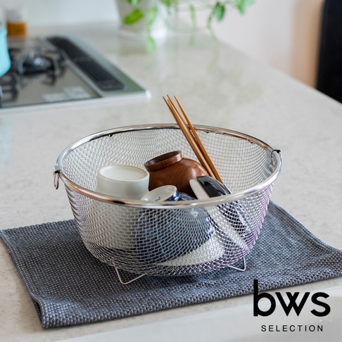 不鏽鋼圓形網狀瀝水籃，內有木碗、白色杯子、藍白碗及筷子，置於灰色布墊上。