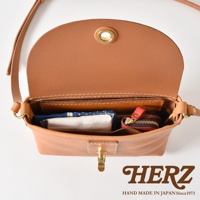 職人手作皮革迷你單肩手袋，採用天然棕色皮革，設計簡約。袋子內裝有幾樣物品，包括紙巾、卡夾和一個紅色零錢包。袋蓋以金屬扣固定，展現細膩的工藝。