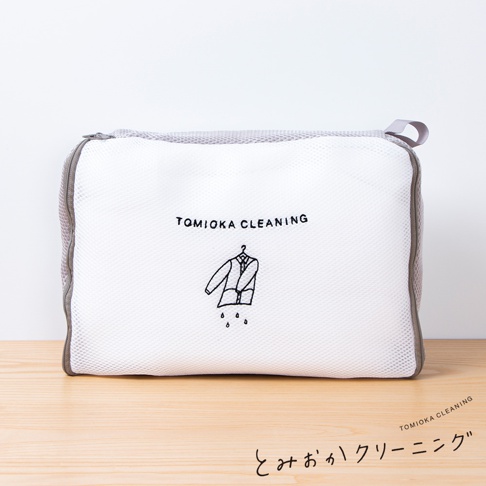 白色網狀方形洗衣袋，邊緣為灰色，印有黑色「TOMIOKA CLEANING」字樣及一件掛在衣架上、滴水的衣物圖案，上方有一個灰色提環。