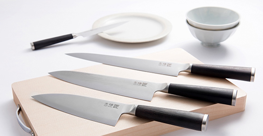 三把銀色刀刃、黑色木柄的萬用刀（Santoku knife）和一把白色小刀，擺放在淺色木製砧板上，背景有白色盤子和碗。