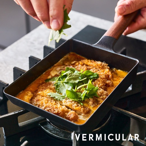 黑色琺瑯鑄鐵玉子燒平底鍋，配有木製手柄，鍋內盛有烹調中的日式炸豬排，上面撒有綠色香草。