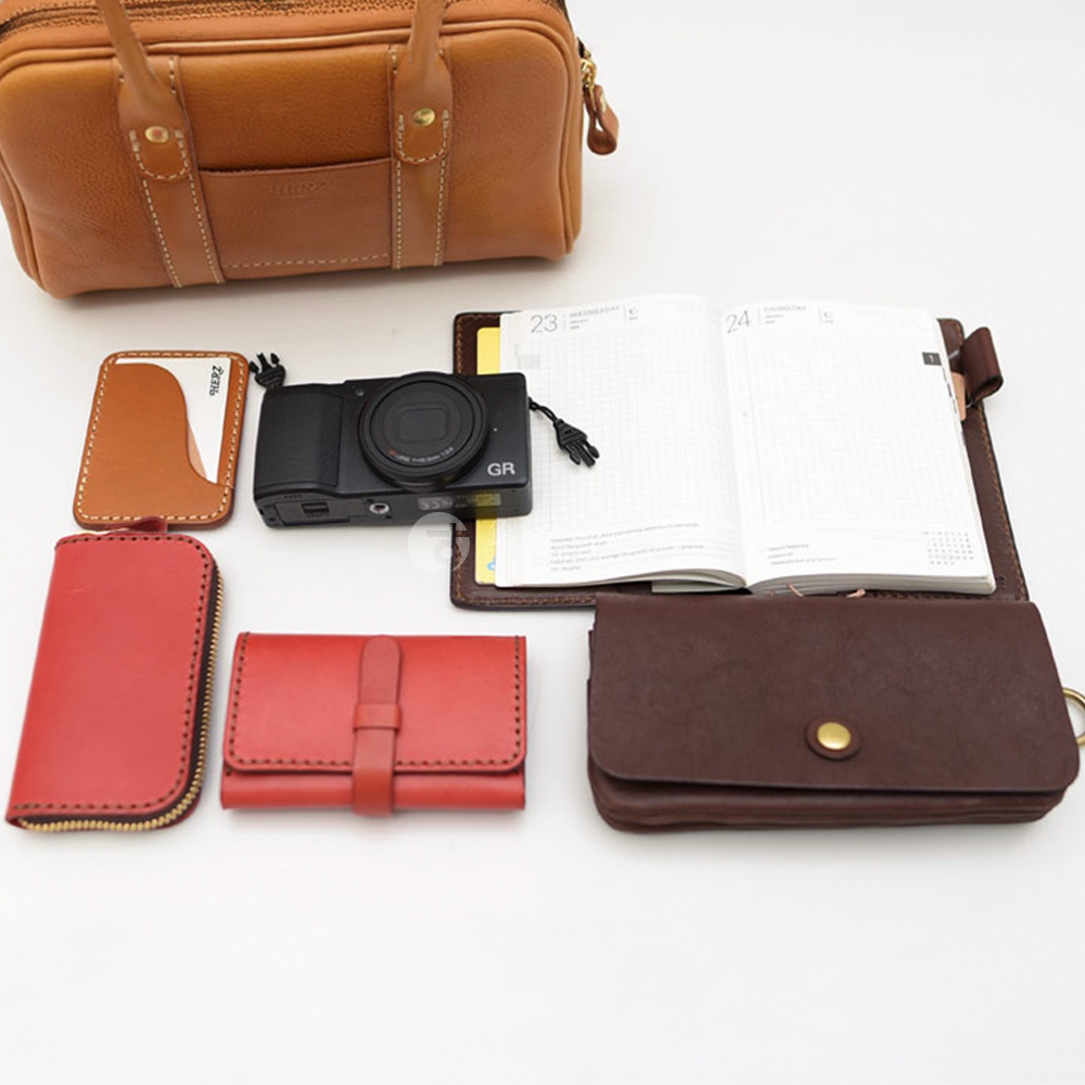 一個皮革波士頓包、一個相機、一本打開的筆記本和幾個皮革配件，呈現在白色背景上。