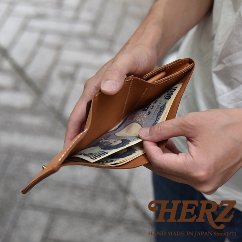 雙手拿著一個日本HERZ職人手作的淺棕色皮革三折迷你短夾，內部夾層展示著幾張一千元面額的日幣紙鈔，整體設計簡約俐落，展現出質感溫潤的皮革細節。