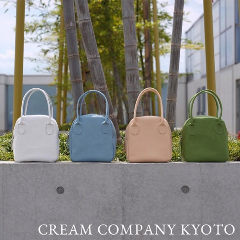 四款皮革手提包，分別為白色、淺藍色、米色和綠色，皆為貝殼造型，配有兩條短提把，置於戶外水泥平台上。