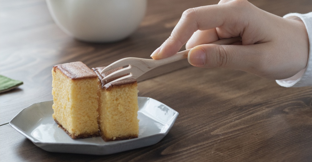 淺色楓木點心叉，具有三叉設計，正在叉取兩塊金黃色、方形的日式蜂蜜蛋糕，蛋糕置於白色六角盤中。