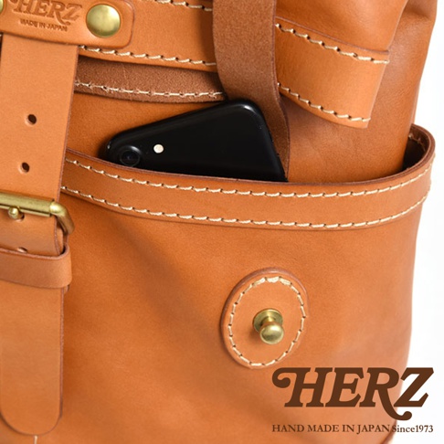 職人手作皮革雙口袋折蓋後背包R-301，特寫展示了焦糖棕色皮革的質感、雙層口袋設計，以及一個置於上方口袋中的黑色手機。包袋採用白色縫線，並有金屬扣環和旋鈕細節。